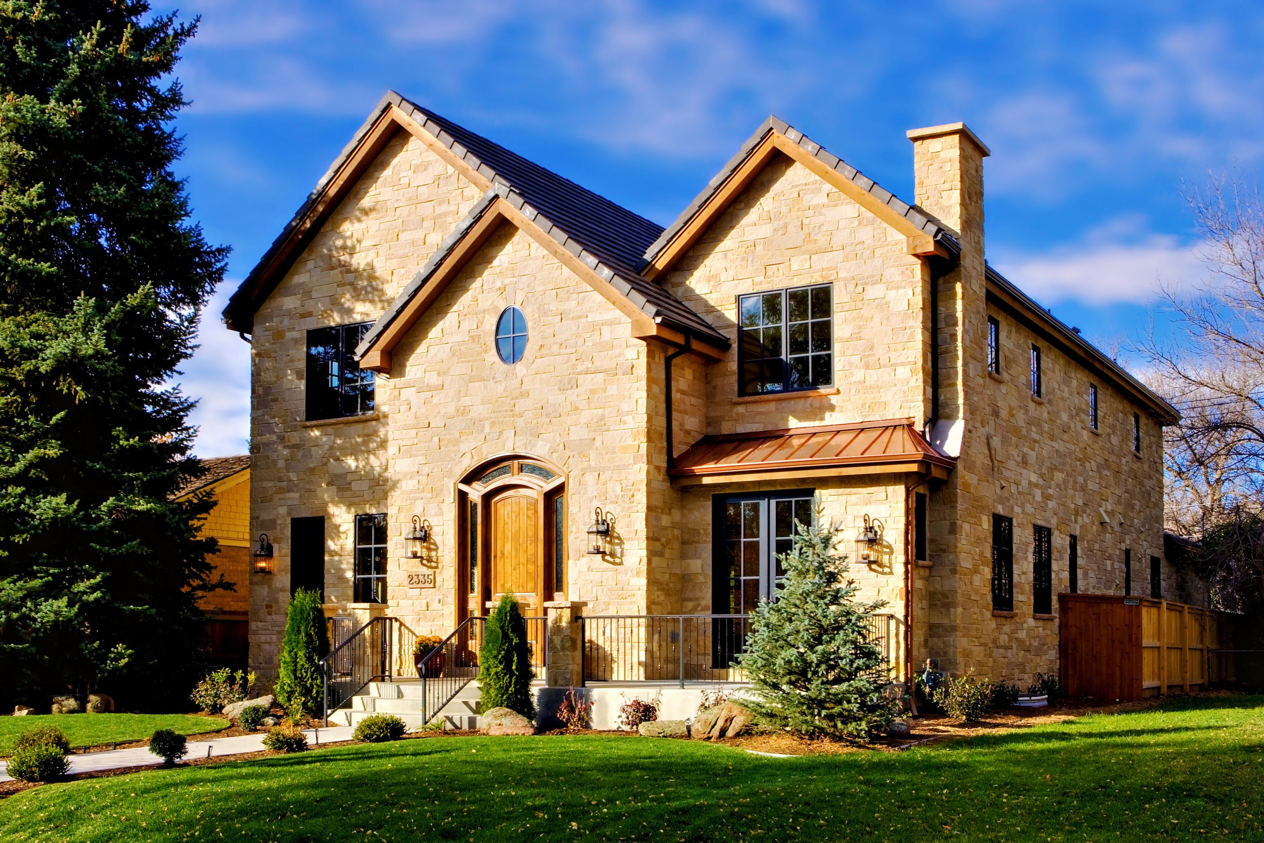 Elegant stone-clad luxury home with arched wooden entryway, manicured lawn, and copper accents in a scenic suburban setting.