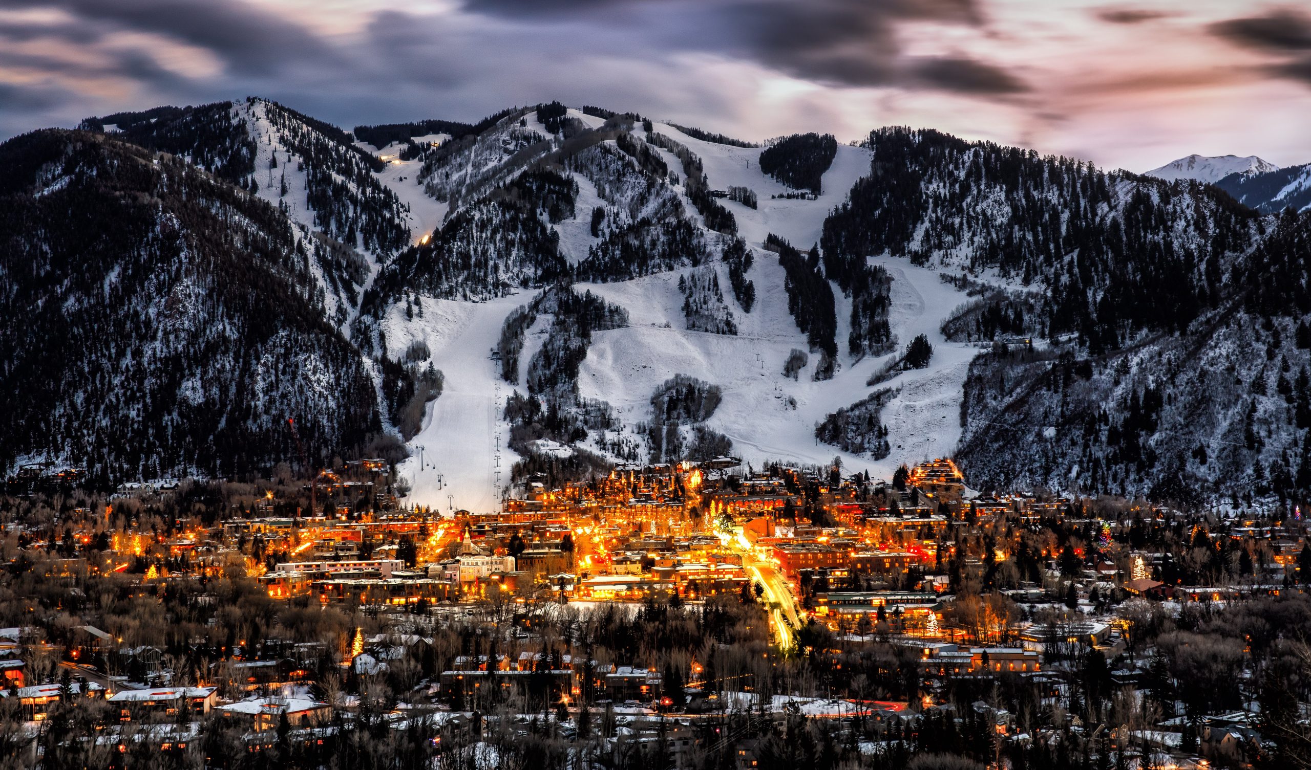 Downtown Aspen Colorado illuminated at dusk with snow-covered Aspen Mountain ski slopes in the background.