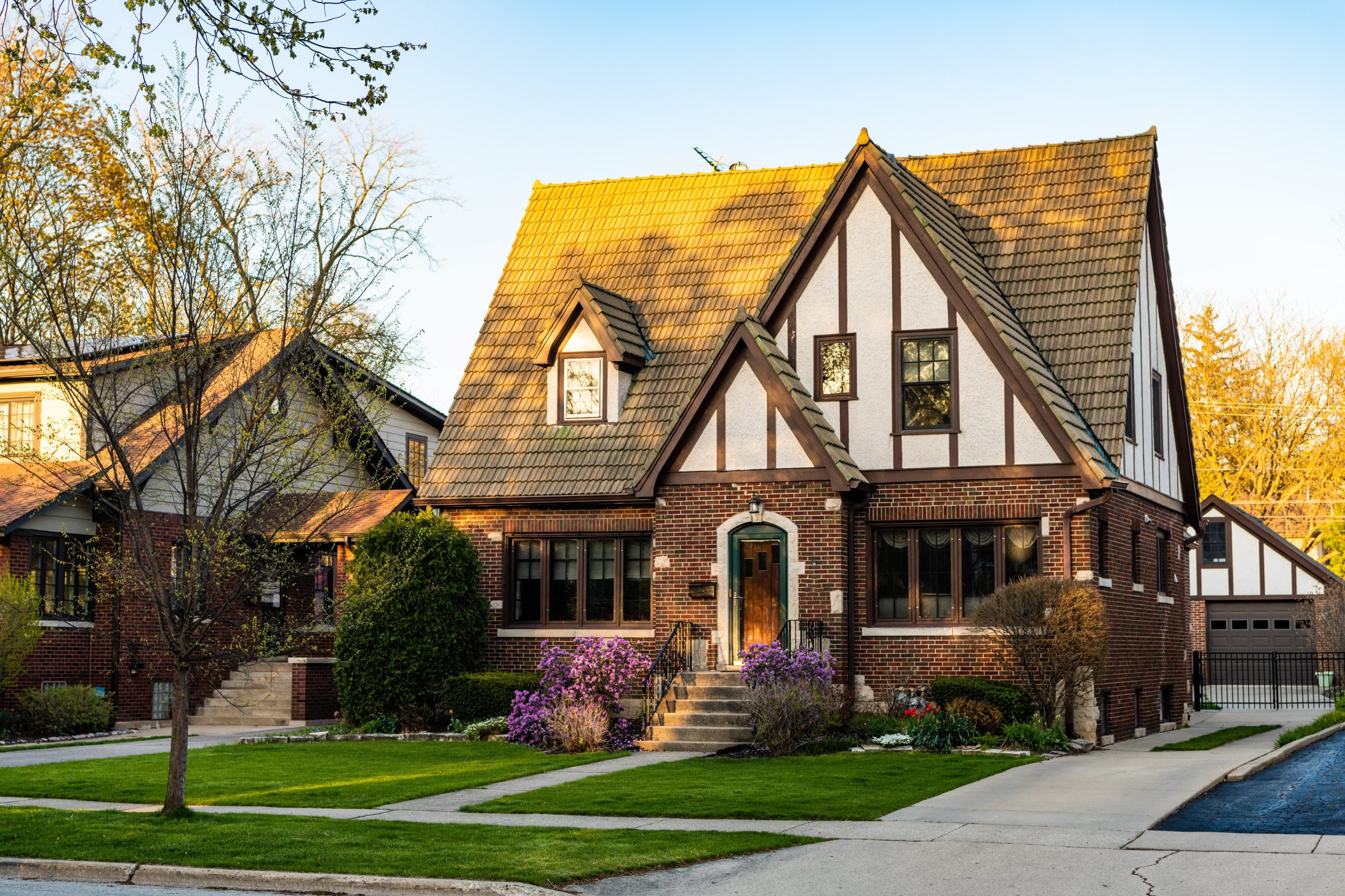Classic Tudor-style home with steep pitched roof, brick and stucco exterior, and lush spring landscaping in a suburban neighborhood.