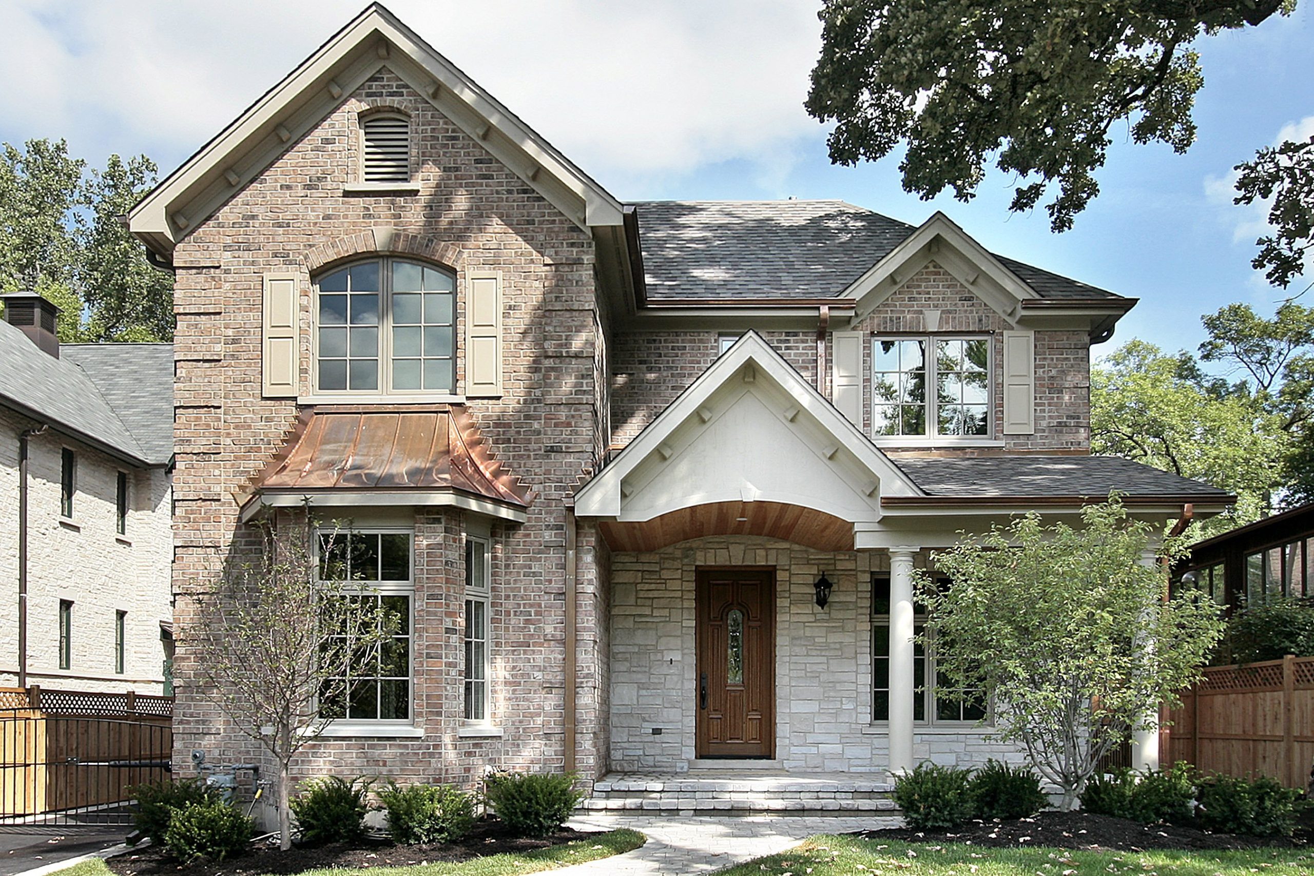 Two-story luxury home with brick and stone exterior, arched copper roof accents, and traditional architectural details in a tree-lined neighborhood.