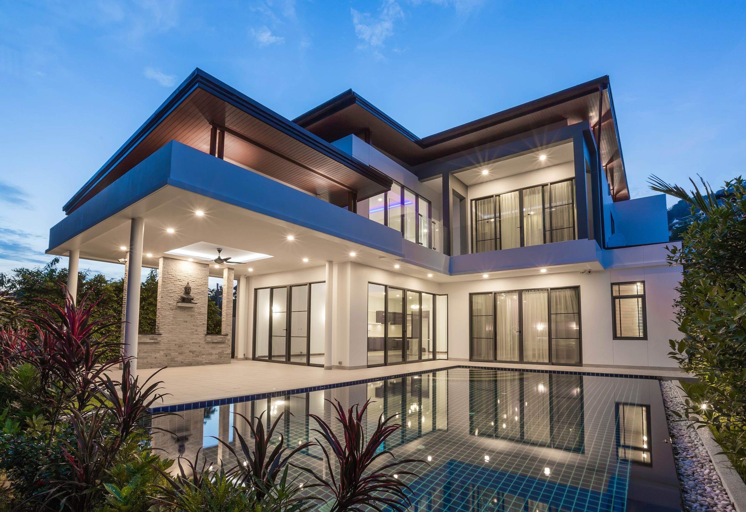 Modern luxury two-story home with expansive glass windows, a covered patio, and a serene reflecting pool at dusk.